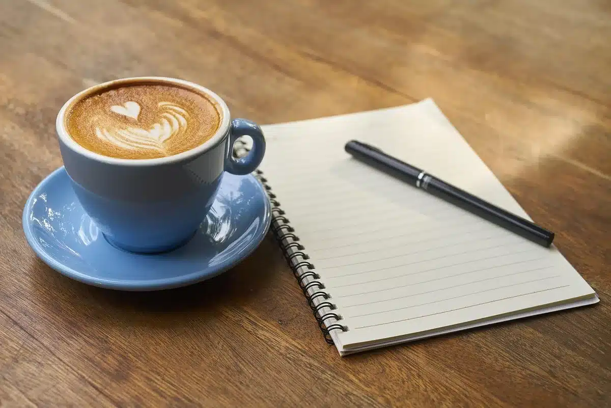 Hands writing a schedule beside a coffee mug on a sunlit desk